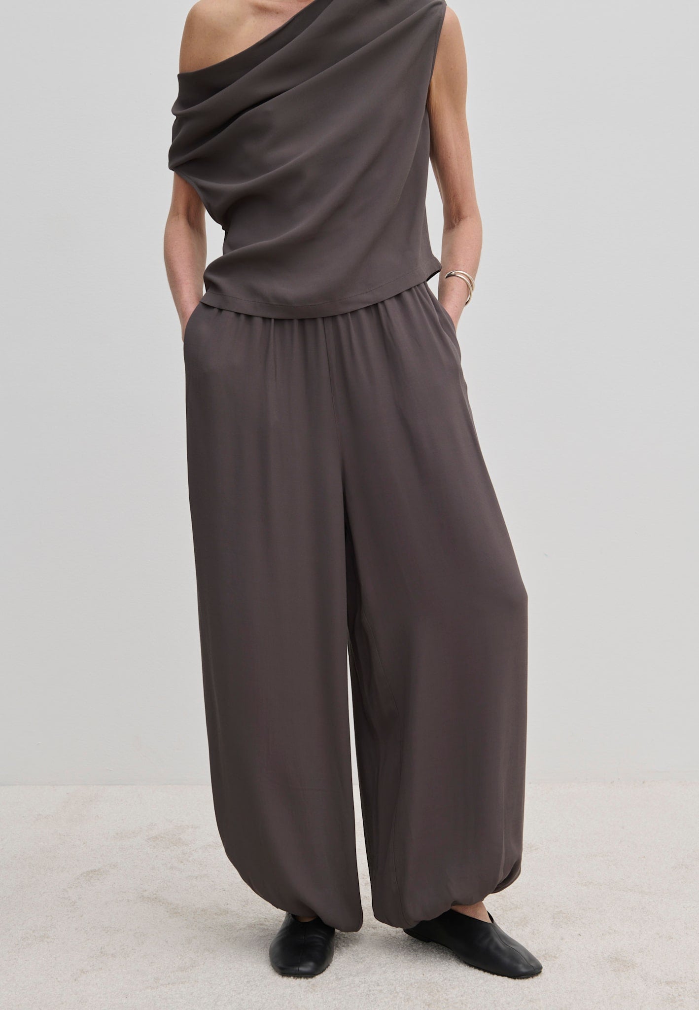 A person wears dark gray Nya Pants by Herskind ApS, featuring balloon legs, an elasticated waist, and a draped top.