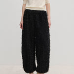 A person wearing a short-sleeved cream top and Herskind ApS Nya Pants—relaxed-fit, voluminous black balloon leg trousers with a textured, fringed surface—paired with cream-colored high heels.