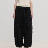 A person wearing a short-sleeved cream top and Herskind ApS Nya Pants—relaxed-fit, voluminous black balloon leg trousers with a textured, fringed surface—paired with cream-colored high heels.