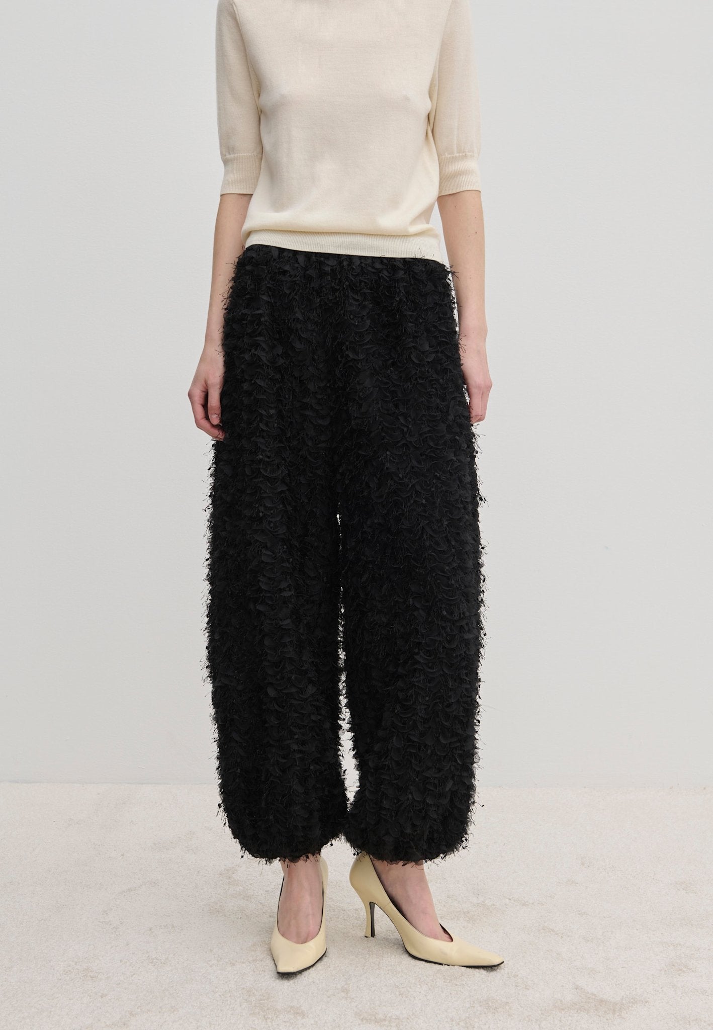 A person wearing a short-sleeved cream top and Herskind ApS Nya Pants—relaxed-fit, voluminous black balloon leg trousers with a textured, fringed surface—paired with cream-colored high heels.