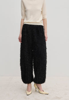 A person wearing a short-sleeved cream top and Herskind ApS Nya Pants—relaxed-fit, voluminous black balloon leg trousers with a textured, fringed surface—paired with cream-colored high heels.