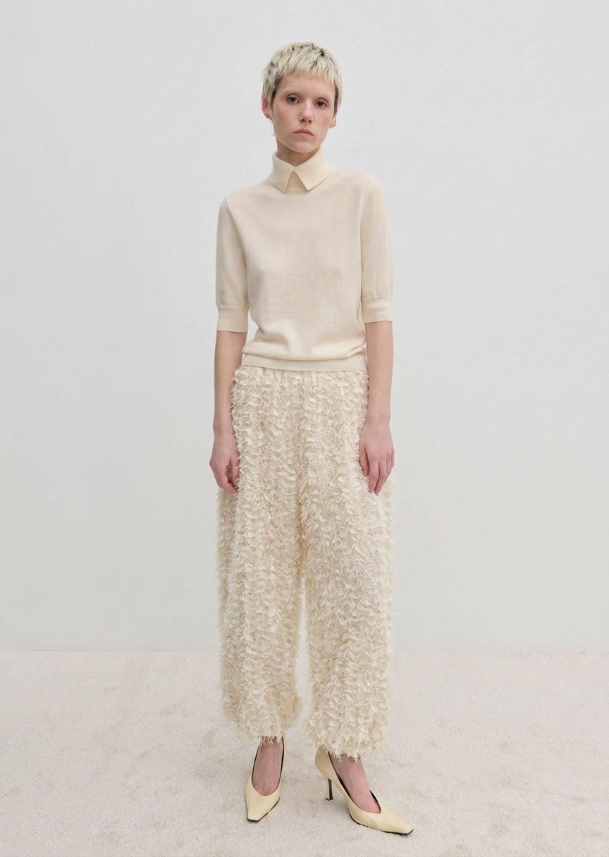A model wearing a short-sleeved cream sweater and Herskind ApS Nya Pants, which are relaxed fit, wide-leg, and textured with a fringed appearance, paired with cream pumps.