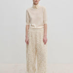 A model wearing a short-sleeved cream sweater and Herskind ApS Nya Pants, which are relaxed fit, wide-leg, and textured with a fringed appearance, paired with cream pumps.