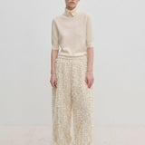 A model wearing a short-sleeved cream sweater and Herskind ApS Nya Pants, which are relaxed fit, wide-leg, and textured with a fringed appearance, paired with cream pumps.