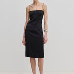 The Amina Dress by Herskind ApS is a knee-length black slip dress with adjustable straps and a square neckline, worn with light-colored heels.