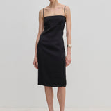 The Amina Dress by Herskind ApS is a knee-length black slip dress with adjustable straps and a square neckline, worn with light-colored heels.