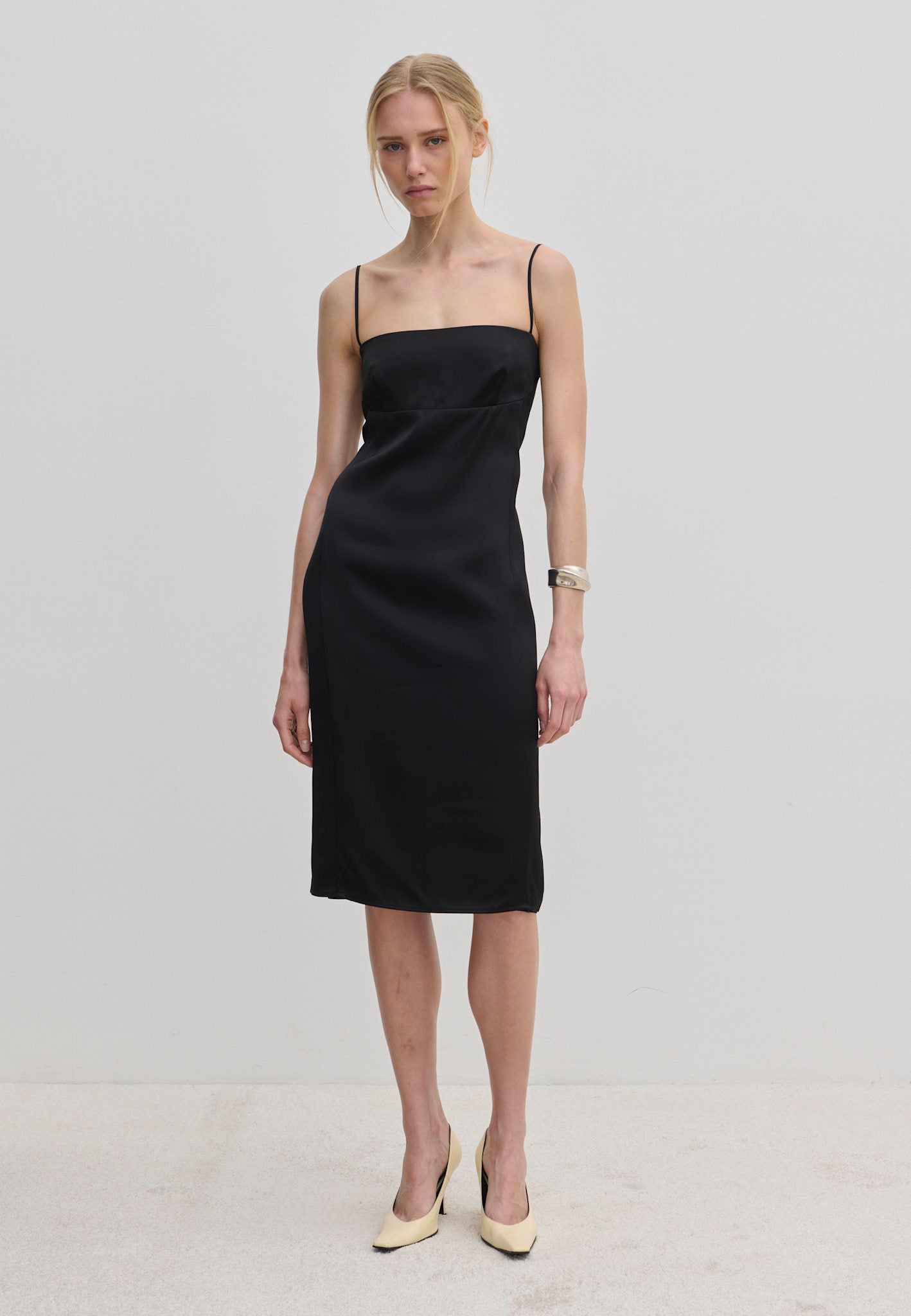 The Amina Dress by Herskind ApS is a knee-length black slip dress with adjustable straps and a square neckline, worn with light-colored heels.