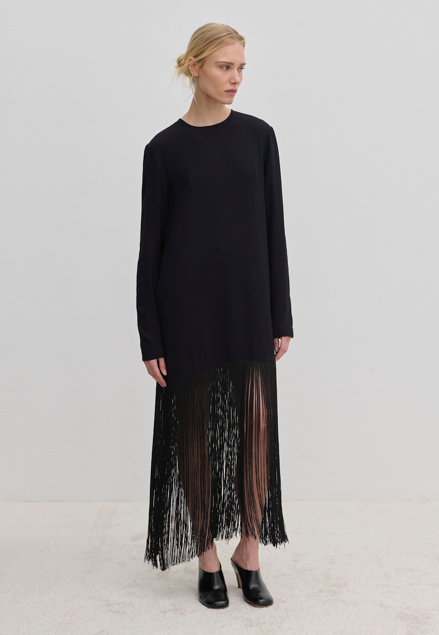 The Aris Dress by Herskind ApS is a long-sleeved black shift dress with a crew neckline and an ankle-length fringe hem, styled with black mules.