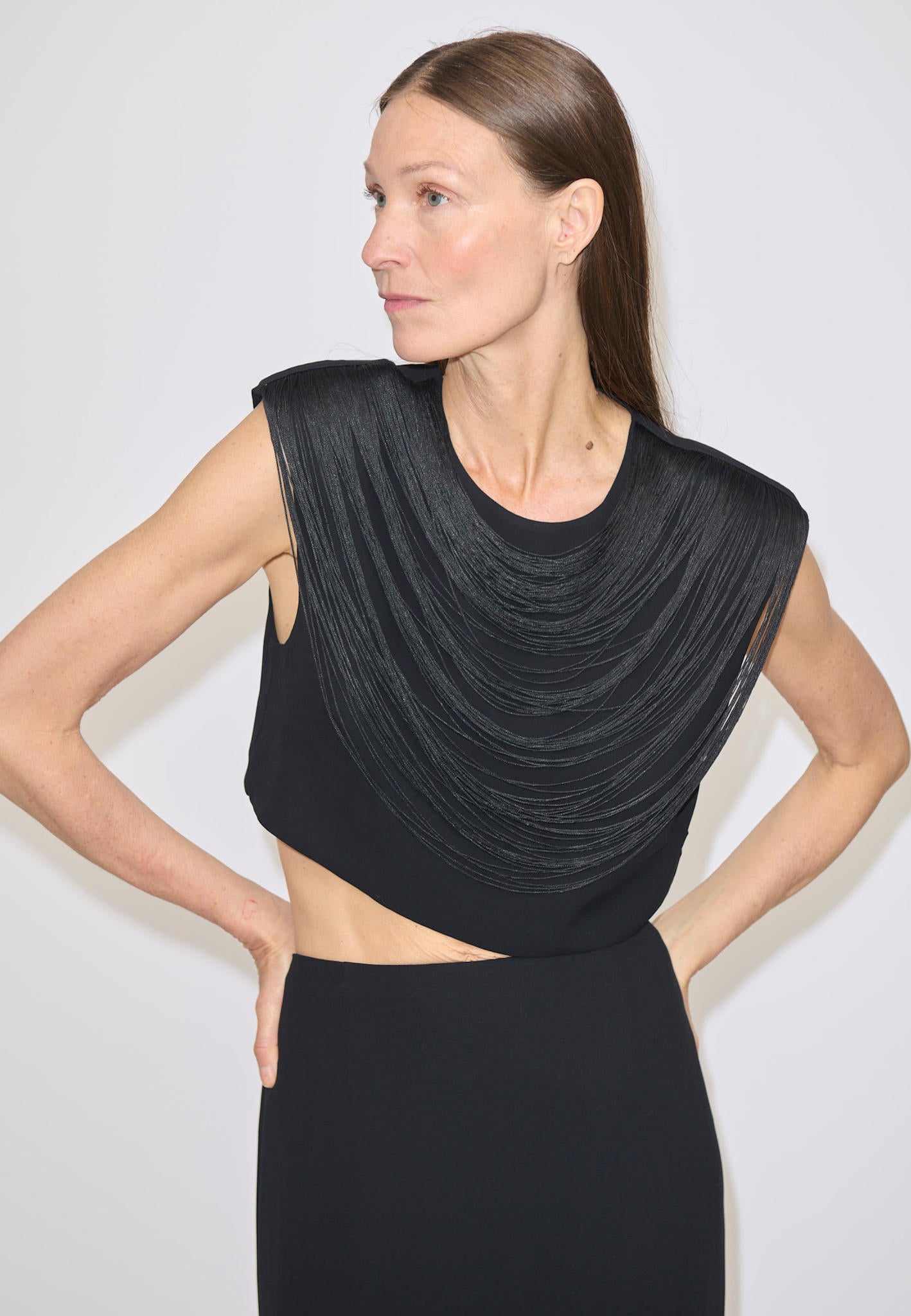 A woman wears a black, cropped Dita Top by Herskind ApS, featuring a sleeveless design with draped fringe layers across the chest and a midriff cut-out.