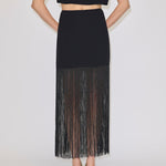 The Herskind ApS Olivia Skirt is a black high-waisted midi skirt with long, sheer fringe at the hem, worn with a matching sleeveless top and cream heels.