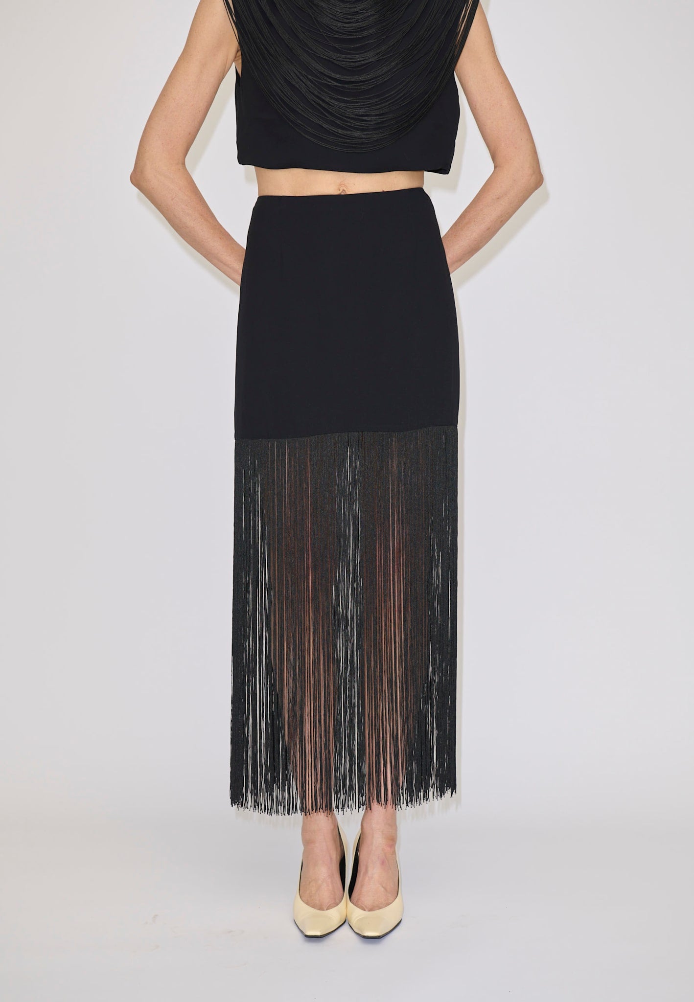 The Herskind ApS Olivia Skirt is a black high-waisted midi skirt with long, sheer fringe at the hem, worn with a matching sleeveless top and cream heels.