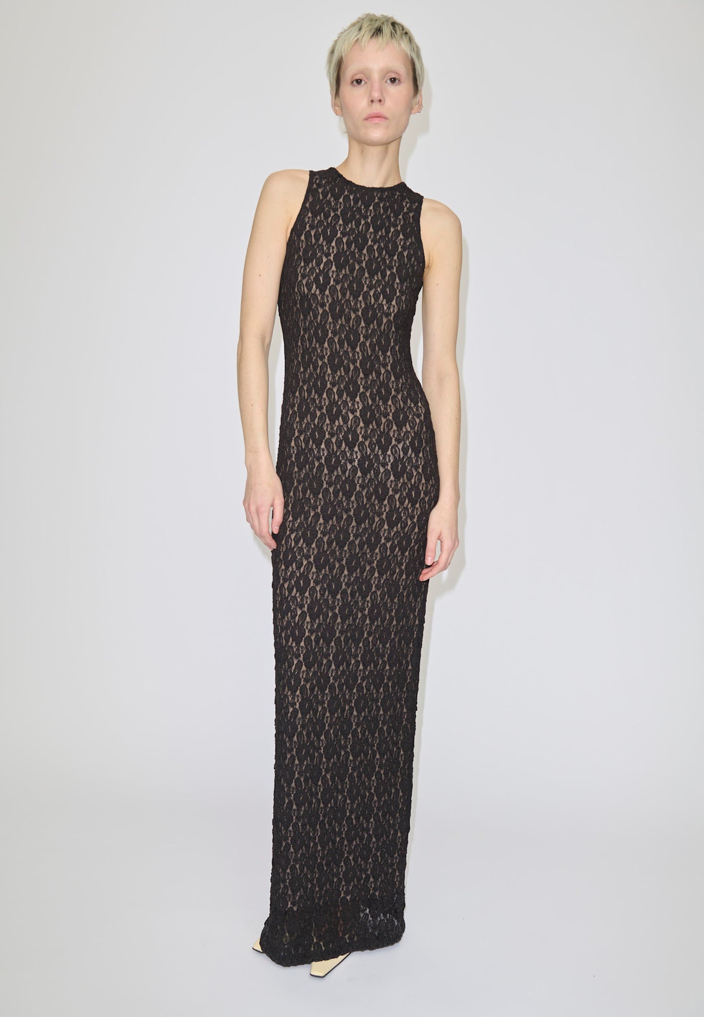 A person wearing the Herskind ApS Chris Dress, a floor-length, sleeveless black lace maxi dress over a nude lining.