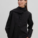 A person wears an oversized black cotton Natalia Shirt by Herskind ApS with an attached scarf-like neck detail and tan trousers.