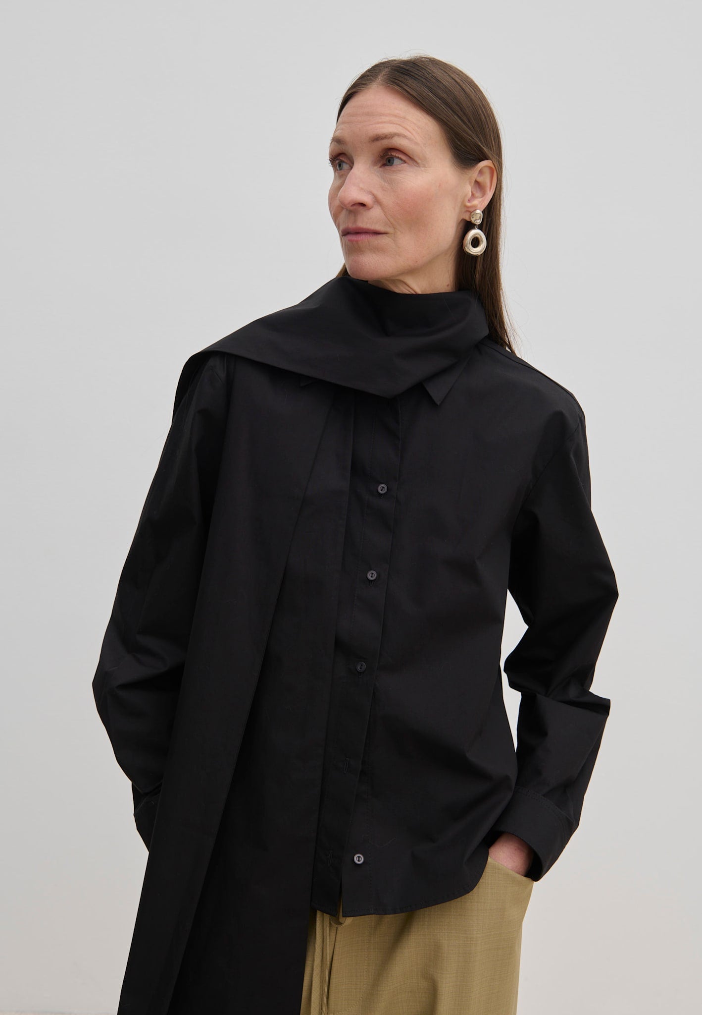 A person wears an oversized black cotton Natalia Shirt by Herskind ApS with an attached scarf-like neck detail and tan trousers.