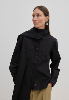 A person wears an oversized black cotton Natalia Shirt by Herskind ApS with an attached scarf-like neck detail and tan trousers.