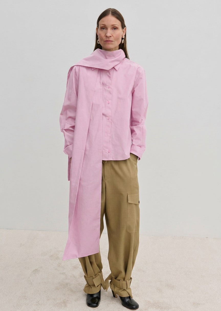 A person wears the Herskind ApS Natalia Shirt, an oversized, long-sleeved light pink button-down with a detachable scarf panel at the shoulder, paired with wide-leg tan cargo pants.