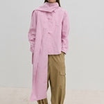 A person wears the Herskind ApS Natalia Shirt, an oversized, long-sleeved light pink button-down with a detachable scarf panel at the shoulder, paired with wide-leg tan cargo pants.