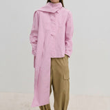 A person wears the Herskind ApS Natalia Shirt, an oversized, long-sleeved light pink button-down with a detachable scarf panel at the shoulder, paired with wide-leg tan cargo pants.