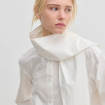 The Natalia Shirt by Herskind ApS is an oversized white cotton button-up with an attached scarf-like collar piece draped around the neck.