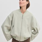 A person wears a pale green, cropped-length Monica Bomber Jacket by Herskind ApS made of wool and alpaca with a ribbed collar and cuffs, paired with dark trousers.