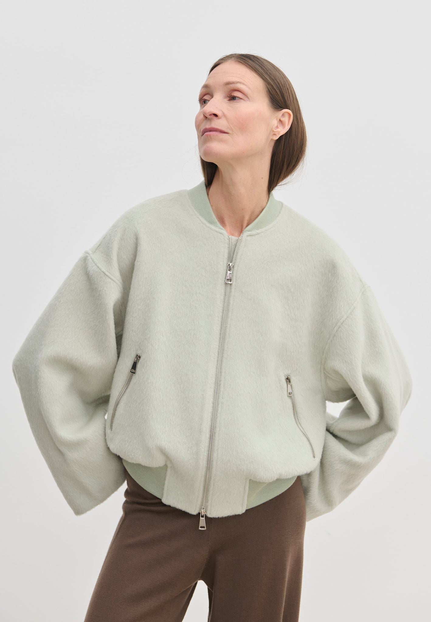 A person wears a pale green, cropped-length Monica Bomber Jacket by Herskind ApS made of wool and alpaca with a ribbed collar and cuffs, paired with dark trousers.