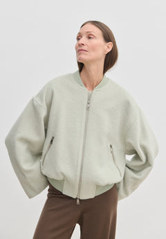 A person wears a pale green, cropped-length Monica Bomber Jacket by Herskind ApS made of wool and alpaca with a ribbed collar and cuffs, paired with dark trousers.