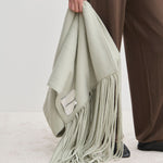 A person holding an oversized, light green Daniella Scarf by Herskind ApS, featuring long fringe.