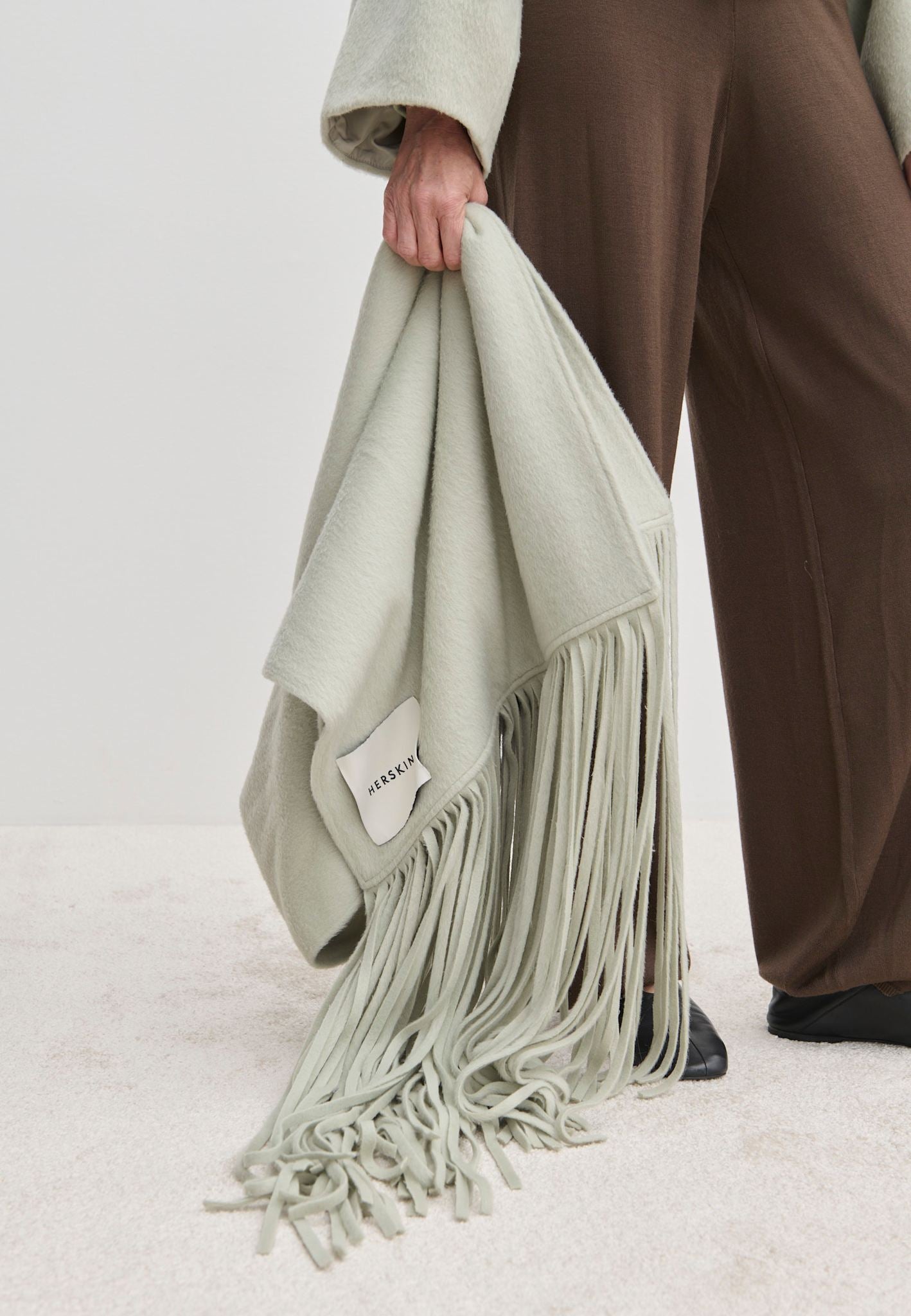 A person holding an oversized, light green Daniella Scarf by Herskind ApS, featuring long fringe.